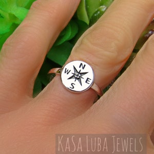 May include: A silver ring featuring a compass design. The ring has a circular face with the cardinal directions (N, S, E, W) and a starburst compass rose. The ring is shown on a finger, with the brand name "KASA LUBA JEWELS" visible.