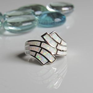 Op de afbeelding: Een zilveren ring met een witte opaal-inleg in een geometrisch patroon. De ring ligt op een witte achtergrond met blauwe glazen stenen.