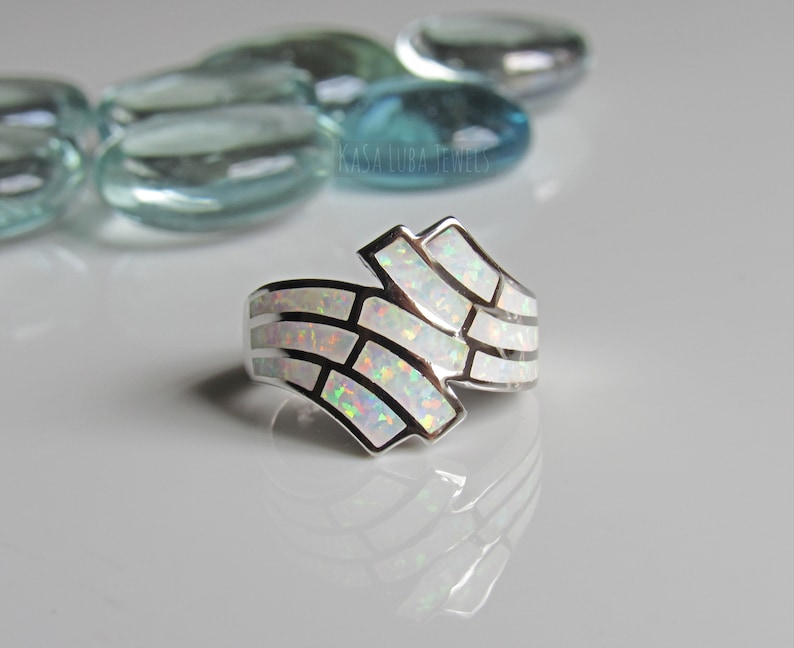Op de afbeelding: Een zilveren ring met een witte opaal-inleg in een geometrisch patroon. De ring ligt op een witte achtergrond met blauwe glazen stenen.
