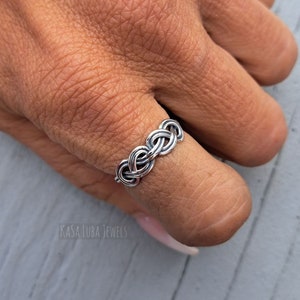 May include: A silver ring with a braided design. The ring is worn on a finger.