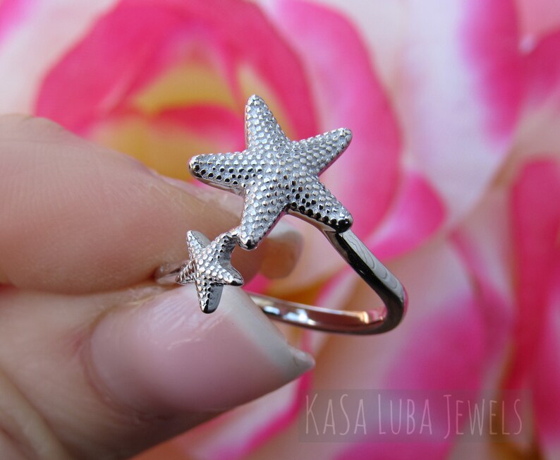 May include: Silver ring with two textured starfish designs. The larger starfish is on top of the smaller starfish. The ring is open and adjustable.