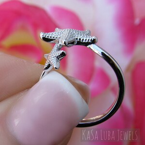 May include: Silver ring with two starfish designs. The ring is open and adjustable. The starfish are facing opposite directions. The ring is on a pink background.
