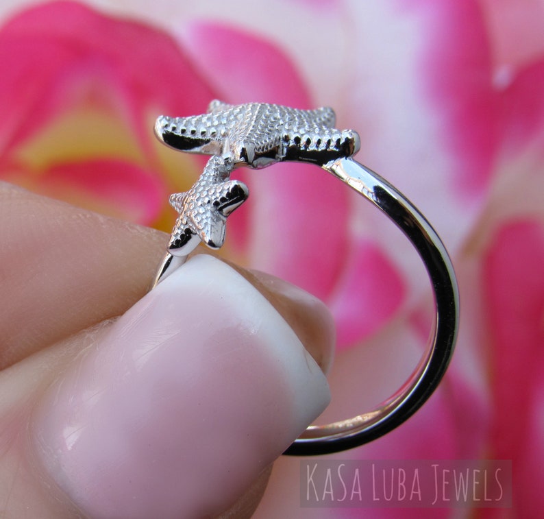 May include: Silver ring with two starfish designs. The ring is open and adjustable. The starfish are facing opposite directions. The ring is on a pink background.