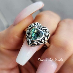May include: A silver ring with a heart-shaped abalone shell inlay. The ring has a detailed, ornate design.