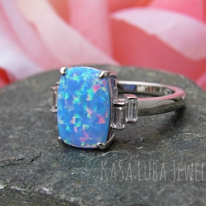May include: A silver ring with a large blue opal gemstone and two smaller clear gemstones on the band. The ring is set on a gray stone surface.