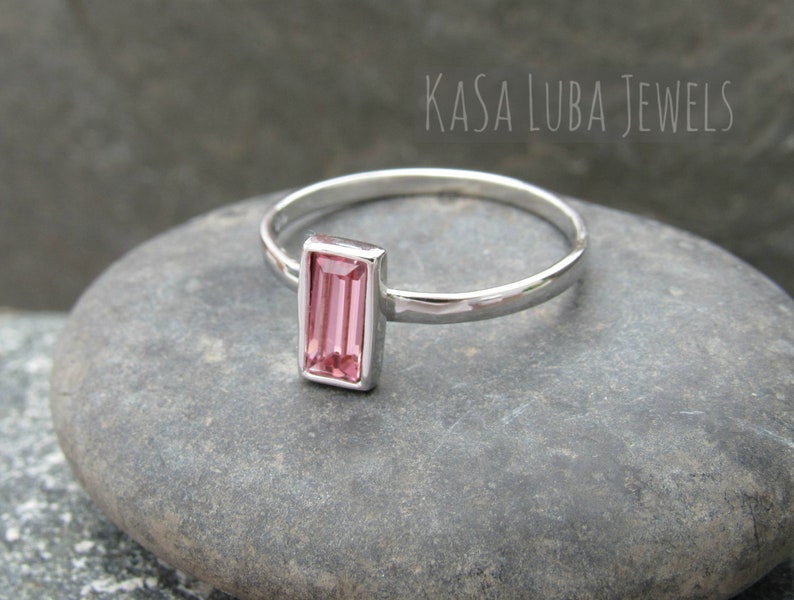 May include: A silver ring with a pink rectangular gemstone. The ring is on a gray stone surface. KASA LUBA JEWELS is written in the background.