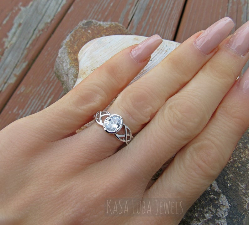 K&ouml;nnte beinhalten: Ein silberner Ring mit einem ovalen, klaren Edelstein. Der Ring hat ein keltisches Knotenmuster auf dem Band. Der Text "KASA LUBA JEWELS" ist unten rechts im Bild zu sehen.