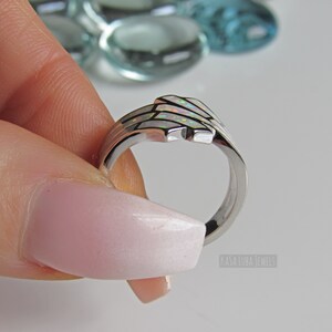 Op de afbeelding: Een zilveren ring met een witte opaal-inleg. De ring is gezet in een delicaat, golvend ontwerp. De ring wordt vastgehouden door een hand met lichtroze nagellak.