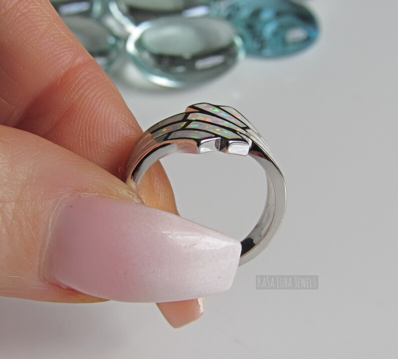 Op de afbeelding: Een zilveren ring met een witte opaal-inleg. De ring is gezet in een delicaat, golvend ontwerp. De ring wordt vastgehouden door een hand met lichtroze nagellak.