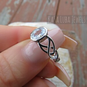 K&ouml;nnte beinhalten: Ein silberner Ring mit einem klaren, ovalen Edelstein. Der Ring hat ein keltisches Knotenmuster am Band und den Text "KASA LUBA JEWELS". Der Ring wird von einer Person mit hellem Hautton gehalten.