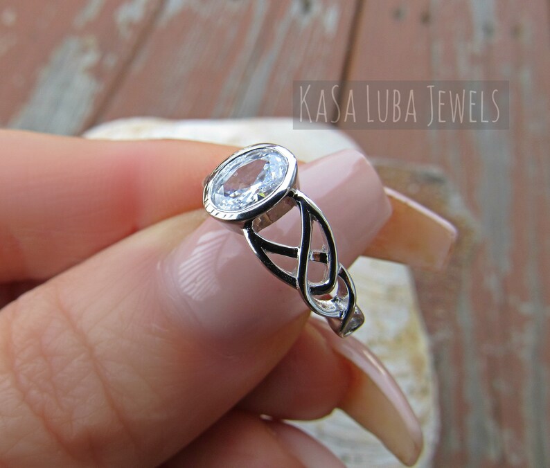 K&ouml;nnte beinhalten: Ein silberner Ring mit einem klaren, ovalen Edelstein. Der Ring hat ein keltisches Knotenmuster am Band und den Text "KASA LUBA JEWELS". Der Ring wird von einer Person mit hellem Hautton gehalten.