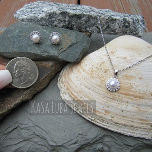 K&ouml;nnte beinhalten: Ein Perlenschmuck-Set mit einer Halskette und Ohrringen. Die Halskette hat eine silberne Kette und einen Anh&auml;nger mit einer Perle, die von einem silbernen Heiligenschein umgeben ist. Die Ohrringe sind silberne Ohrstecker mit einer Perle, die von einem silbernen Heiligenschein umgeben ist. Der Schmuck wird auf einer Muschel und Steinen pr&auml;sentiert.