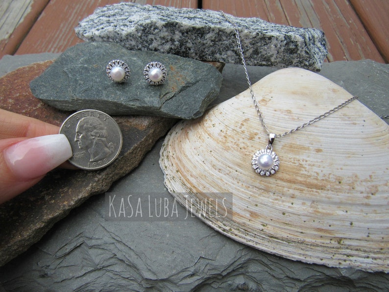 K&ouml;nnte beinhalten: Ein Perlenschmuck-Set mit einer Halskette und Ohrringen. Die Halskette hat eine silberne Kette und einen Anh&auml;nger mit einer Perle, die von einem silbernen Heiligenschein umgeben ist. Die Ohrringe sind silberne Ohrstecker mit einer Perle, die von einem silbernen Heiligenschein umgeben ist. Der Schmuck wird auf einer Muschel und Steinen pr&auml;sentiert.