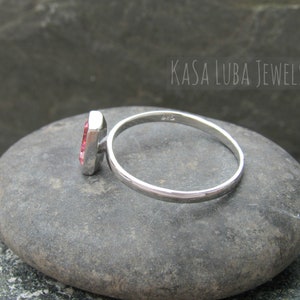May include: A silver ring with a pink gemstone set in a square shape. The ring is on a gray stone.