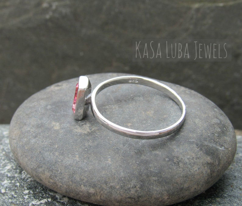 May include: A silver ring with a pink gemstone set in a square shape. The ring is on a gray stone.