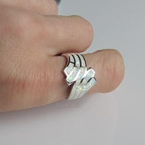 Op de afbeelding: Een zilveren ring met een witte opaal-steen gezet in een geometrisch ontwerp. De ring wordt gedragen aan een vinger.