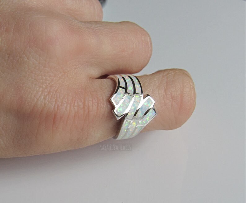 Op de afbeelding: Een zilveren ring met een witte opaal-steen gezet in een geometrisch ontwerp. De ring wordt gedragen aan een vinger.