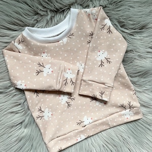 Könnte beinhalten: Ein beiger Langarmsweater mit einem weißen gerippten Ausschnitt und einem Rentier-Druck. Die Rentiere sind weiß mit braunen Geweihen und roten Nasen.