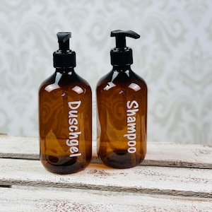 May include: Two amber glass bottles with black pump tops and white labels. The bottle on the left is labeled "Duschgel" and the bottle on the right is labeled "Shampoo".