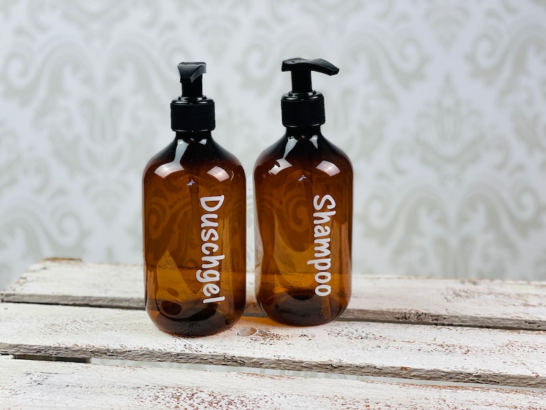 May include: Two amber glass bottles with black pump tops and white labels. The bottle on the left is labeled "Duschgel" and the bottle on the right is labeled "Shampoo".