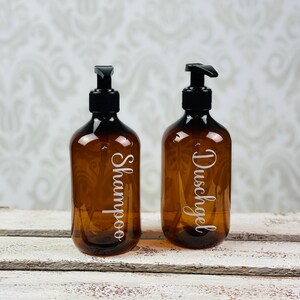 May include: Two amber glass bottles with black pump tops and white labels. The bottle on the left is labeled "Shampoo" and the bottle on the right is labeled "Duschgel".