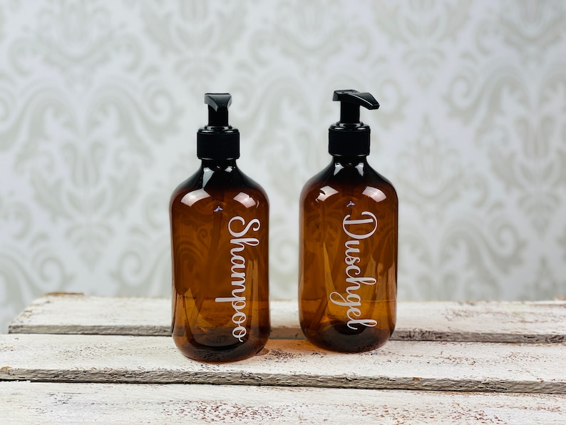 May include: Two amber glass bottles with black pump tops and white labels. The bottle on the left is labeled "Shampoo" and the bottle on the right is labeled "Duschgel".
