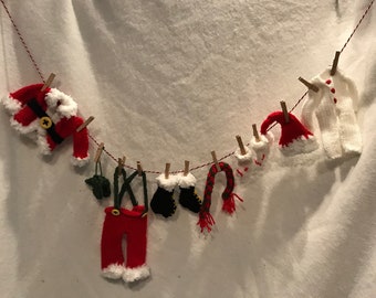 May include: A string of miniature knitted Christmas clothing items hanging on a clothesline. The items include a Santa suit, a pair of pants, a hat, mittens, a scarf, and a white onesie.