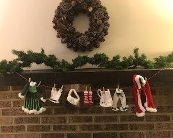 May include: A string of miniature clothing items, including a green dress, a white dress, a white shirt, a pair of red and white striped stockings, a white dress with a red bow, a pair of blue pants, and a red and white Santa suit, are hanging on a clothesline against a brick wall.