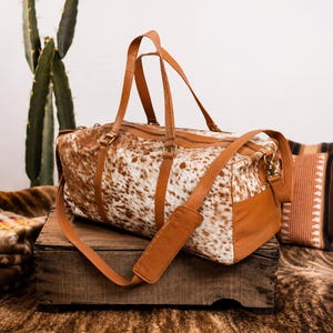 May include: A duffel bag featuring a white and brown cowhide pattern on the main body, complemented by tan leather handles, side panels, and an adjustable shoulder strap with a padded section. The bag includes a top zipper closure and metal hardware.
