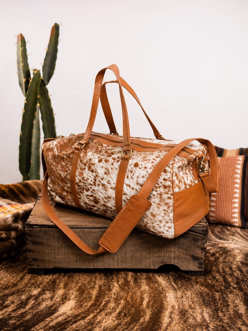May include: A duffel bag featuring a white and brown cowhide pattern on the main body, complemented by tan leather handles, side panels, and an adjustable shoulder strap with a padded section. The bag includes a top zipper closure and metal hardware.