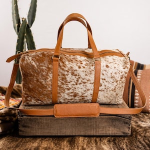 May include: A duffel bag crafted from brown and white cowhide featuring tan leather handles, a matching adjustable shoulder strap with a padded section, and a top zipper closure. The bag has a rectangular shape with reinforced leather trim.