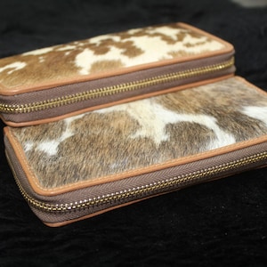 Puede incluir: Dos carteras de piel de vaca marr&oacute;n y blanca con cremalleras doradas. Las carteras est&aacute;n apiladas una encima de la otra.