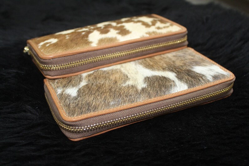 Puede incluir: Dos carteras de piel de vaca marr&oacute;n y blanca con cremalleras doradas. Las carteras est&aacute;n apiladas una encima de la otra.