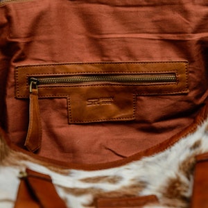 May include: The interior of a bag featuring a brown fabric lining and a leather pocket with a brass zipper. A leather patch is embossed with the text SR SHIRAZ RUGS. The exterior of the bag is made of white and brown cowhide with brown leather straps.