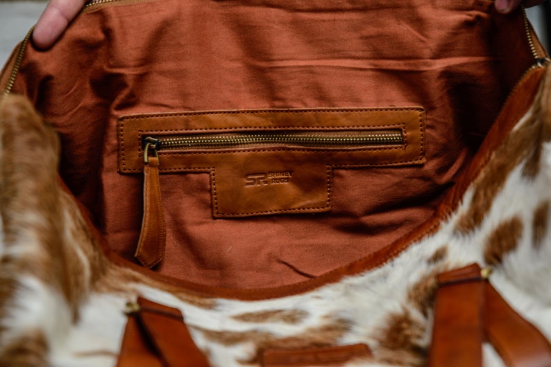 May include: The interior of a bag featuring a brown fabric lining and a leather pocket with a brass zipper. A leather patch is embossed with the text SR SHIRAZ RUGS. The exterior of the bag is made of white and brown cowhide with brown leather straps.