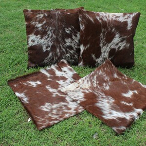 May include: Four brown and white cowhide throw pillows with a natural, spotted pattern. The pillows are arranged on a green grassy surface.