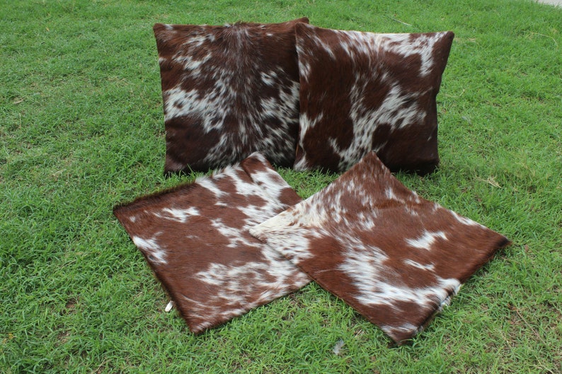 May include: Four brown and white cowhide throw pillows with a natural, spotted pattern. The pillows are arranged on a green grassy surface.