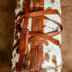 May include: A duffel bag crafted from white and brown cowhide with a textured fur finish. The bag features smooth brown leather trim, a central zipper closure, two top handles, and an adjustable shoulder strap with a padded leather section.