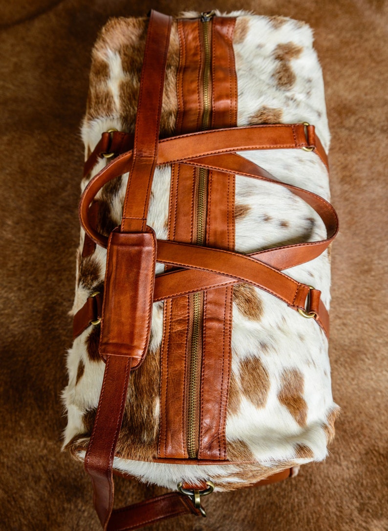 May include: A duffel bag crafted from white and brown cowhide with a textured fur finish. The bag features smooth brown leather trim, a central zipper closure, two top handles, and an adjustable shoulder strap with a padded leather section.