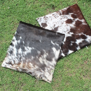 May include: Two cowhide throw pillows with a black and white pattern. The pillow on the left has a brown undertone, while the pillow on the right has a reddish-brown undertone.