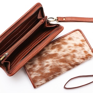 May include: A brown leather wallet with a zipper closure and a matching wrist strap. The wallet is open to reveal multiple card slots and a bill compartment. The wallet is made of cowhide with a brown and white pattern.