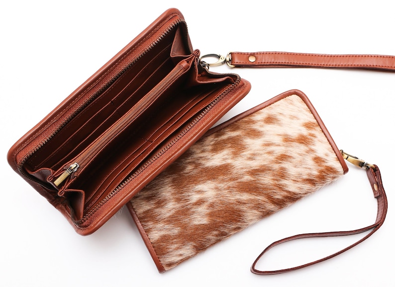 May include: A brown leather wallet with a zipper closure and a matching wrist strap. The wallet is open to reveal multiple card slots and a bill compartment. The wallet is made of cowhide with a brown and white pattern.