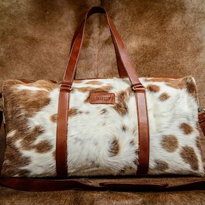May include: A rectangular duffel bag featuring a white and brown cowhide pattern with a textured hair-on finish. The bag includes brown leather handles, a matching adjustable shoulder strap, and a small rectangular leather patch on the front.