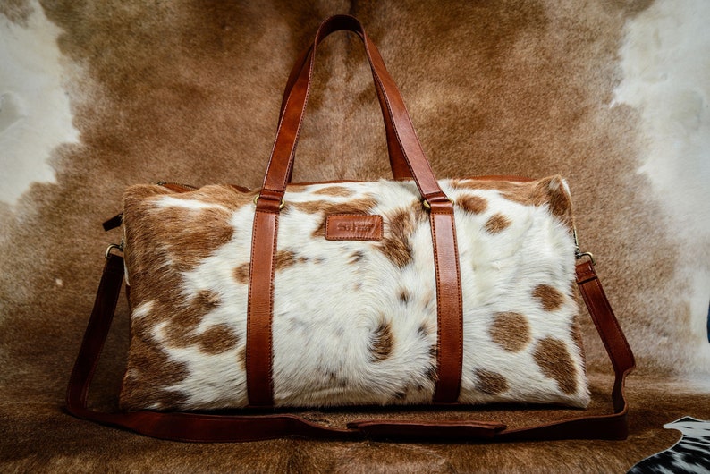 May include: A rectangular duffel bag featuring a white and brown cowhide pattern with a textured hair-on finish. The bag includes brown leather handles, a matching adjustable shoulder strap, and a small rectangular leather patch on the front.