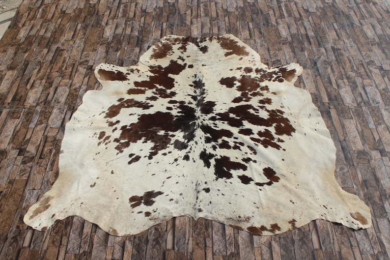 May include: A white cowhide rug with brown and black spots. The rug is laid out on a brown tiled floor.