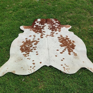 May include: A white cowhide rug with brown spots. The rug is laid out on a green grassy surface.