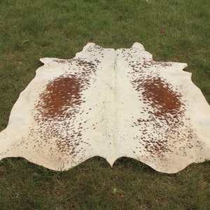 May include: A white cowhide rug with brown spots. The rug is laid out on a green grassy surface.