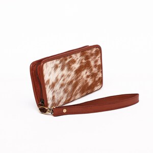 May include: A brown leather wallet with a white and brown cowhide pattern. The wallet has a zipper closure and a brown leather strap.
