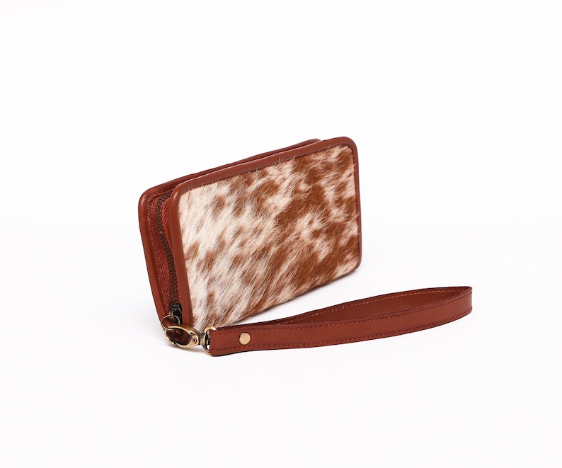 May include: A brown leather wallet with a white and brown cowhide pattern. The wallet has a zipper closure and a brown leather strap.
