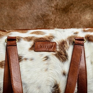 May include: A duffel bag featuring a white and brown cowhide pattern with smooth brown leather handles and a rectangular leather patch embossed with the text ST. SIMON BAG. The bag includes gold-toned metal hardware connecting the straps.